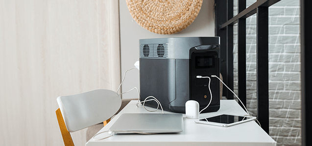 un regulador de voltaje de color gris con negro se encuentra sobre una mesa de color blanco pegada a una ventana mientras se están cargando una laptop y una tablet