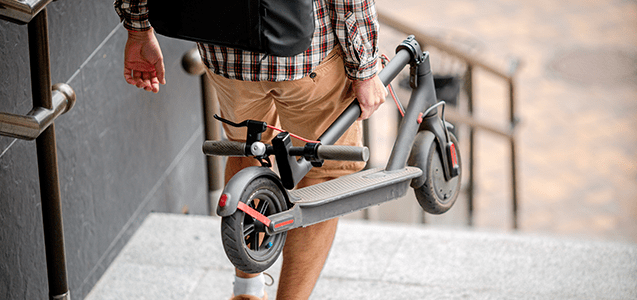 un estudiante de genero masculino se encuentra trasladando a pie su scooter electrico plegable mientras baja las escaleras